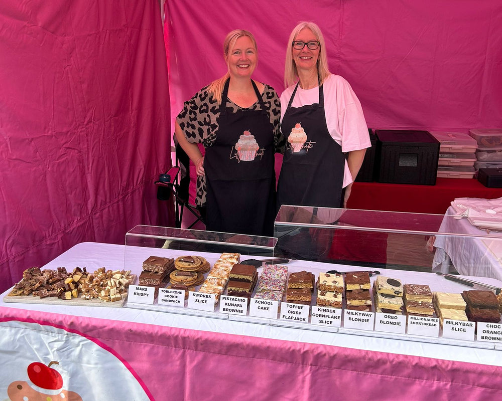 Two women behind a cake stand with various cakes at a pink tented event.